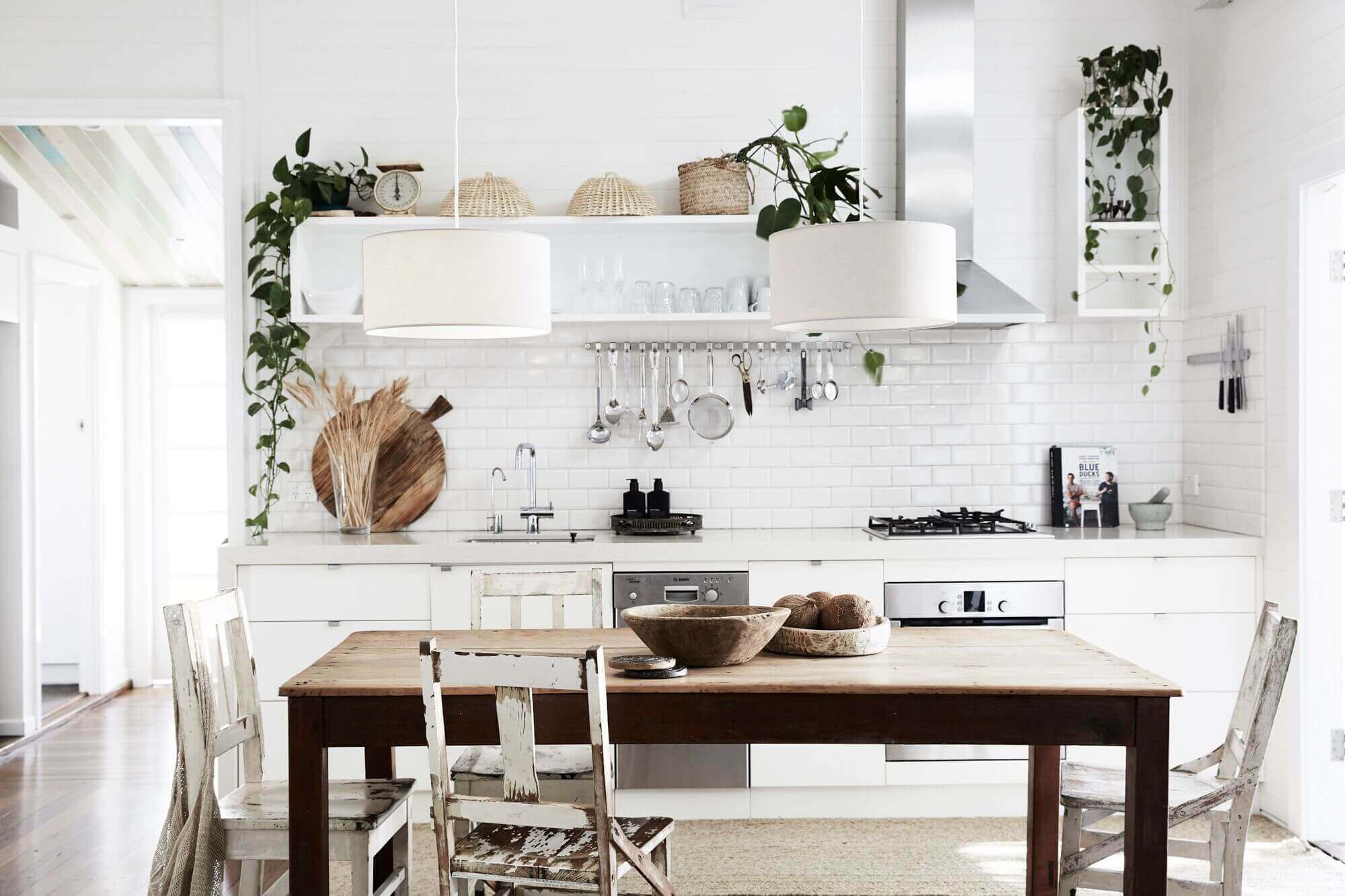 The Cottage, Byron Beach Abodes: Open plan kitchen with white subway tiles, rustic timber table, mixed chairs, open shelves and trailling greenery